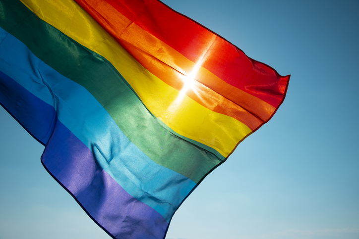 closeup of a rainbow flag waving on the blue sky, moved by the wind, with the sun in the background