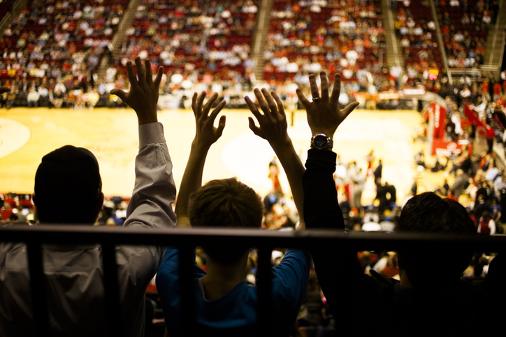 Unrecognizable+people+raise+hands+in+excitement+during+a+basketball+sports+game+inside+a+large+basketball+sports+stadium.+Basketball+court+seen+below.+Silhouette.+Cheering+fans.