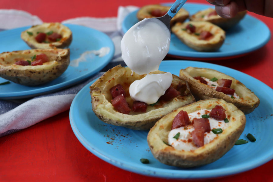 containing a group of crispy, baked potato skins loaded with melted cheese, bacon cubes and sour cream, garnished with a sprinkling of chopped chives,  sitting beside blue and white stripped tea towel on red surface.