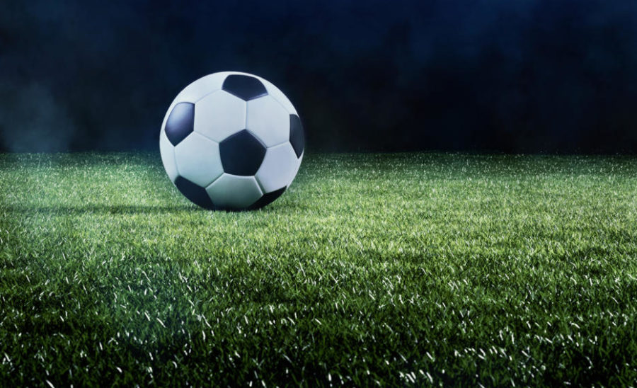 Football or soccer background with a row of spotlights illuminating a ball on the green turf in a stadium in a sports championship or World Cup event in a panorama banner