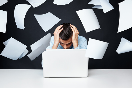 Stressed man sitting by laptop while papers fall over him.