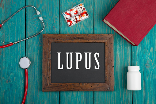 Blackboard with text "Lupus", book, pills and stethoscope on blue wooden background.