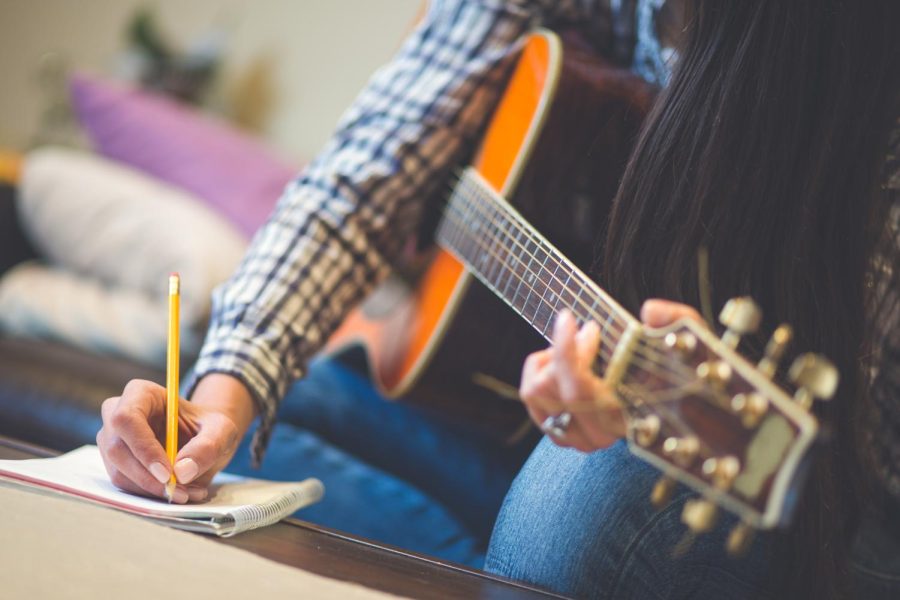 A woman writes on a notepad as she holds a guitar. Writing your own lyrics makes you an artist. Singing someone elses lyrics makes you a performer. 