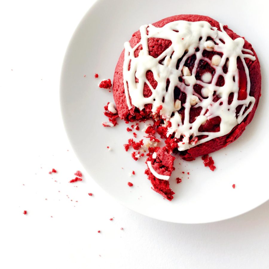 A red velvet cookie with white frosting with a chunk broken off and crumbles on the table. 
