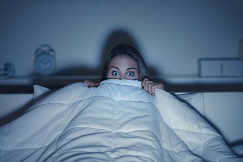A young woman hiding under the covers as the light from a television lights her up.