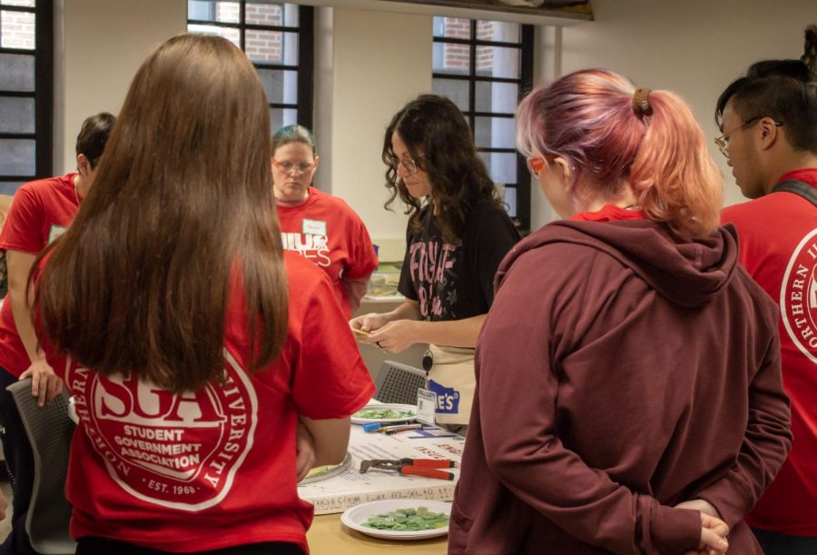 Danielle Casali, an artist, muralist and St. Charles resident, shows volunteers how to cut materials for the City of DeKalb Mosaic at the DeKalb Public Library. The library was one of the six volunteer locations for NIU Cares, an annual day of service. (Madelaine Vikse | Northern Star) 