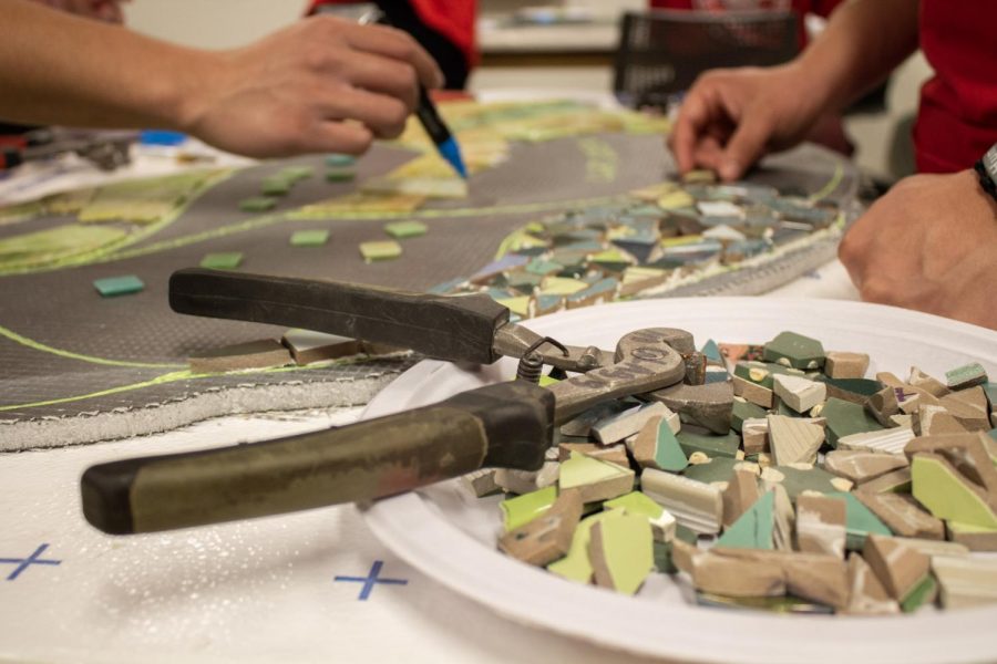 A plate holds shards of stained glass and ceramic for volunteers to cut and arrange sits on a table while they work to lay the tesserae on one of the displays. (Madelaine Vikse | Northern Star) 