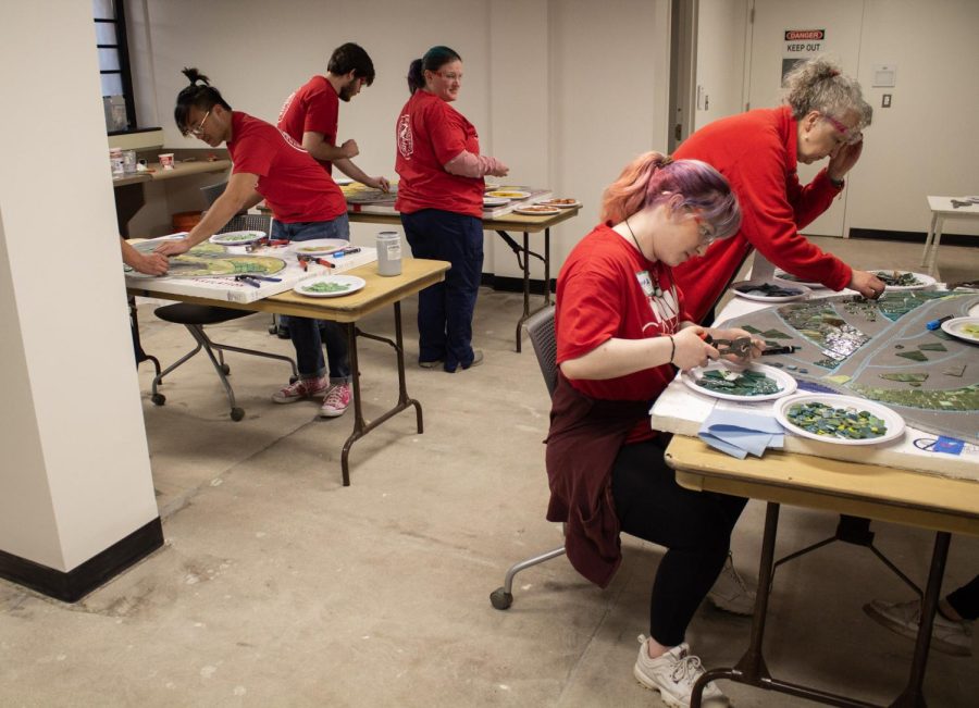 Volunteers cut materials and attach them to different displays with environmental themes. One of the volunteering locations featured work on a City of DeKalb mosaic mural at the DeKalb Public Library, 309 Oak Street. (Madelaine Vikse | Northern Star) 