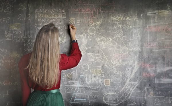 A woman in a red long-sleeve writes on a chalkboard. While advanced placement courses sound beneficial, dual-credit courses will better prepare students for college. (Andrea Piacquadio | Pexels)