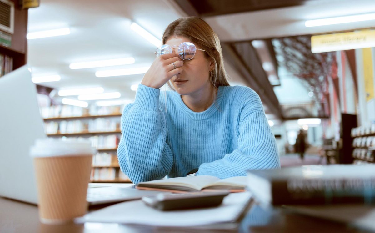 A woman rubs her eyes as she sits a a library table covered in books. Professors should be more understanding toward students during midterm week. (Courtesy of Getty Images)