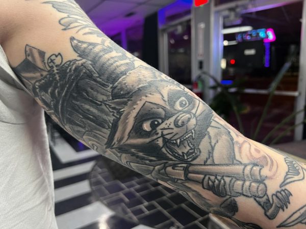 A tattoo on Proton Tattoo’s tattoo artist Grant Katula’s arm shows a raccoon guarding a trash can with a shotgun. Students shared their first tattoo stories and advise others to consider the price and art that’ll be imprinted on their bodies. (Brandon Clark | Northern Star)