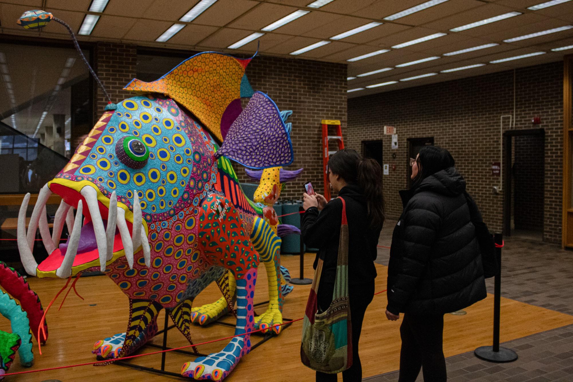 Perla Marrufo (left), a junior psychology major, takes a photo of the alebrijes “Patapez” art installation while Ajla Omerovic, a senior accounting major, admires the sculpture. The sculpture’s bright colors and patterns caught the students’ eyes while walking through the lobby at the Founder Memorial Library. (Totus Tuus Keely | Northern Star)