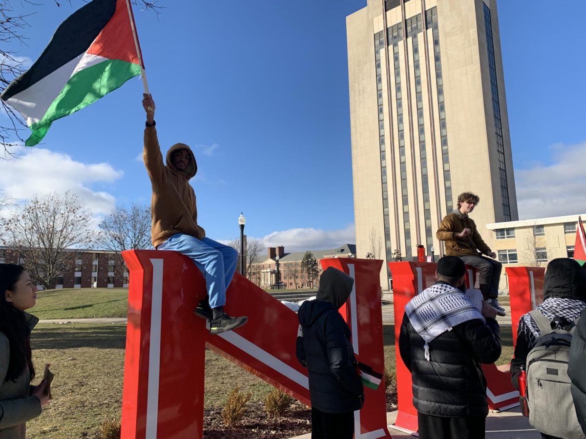 First-year history major Michael Ingram waves the Palestinian flag with one hand while he sits on top of the “N” of the “Huskie Pride” sculpture in MLK Commons. Students protested in support of Palestine for the second consecutive week last Friday in MLK Commons. (Rachel Cormier | Northern Star)