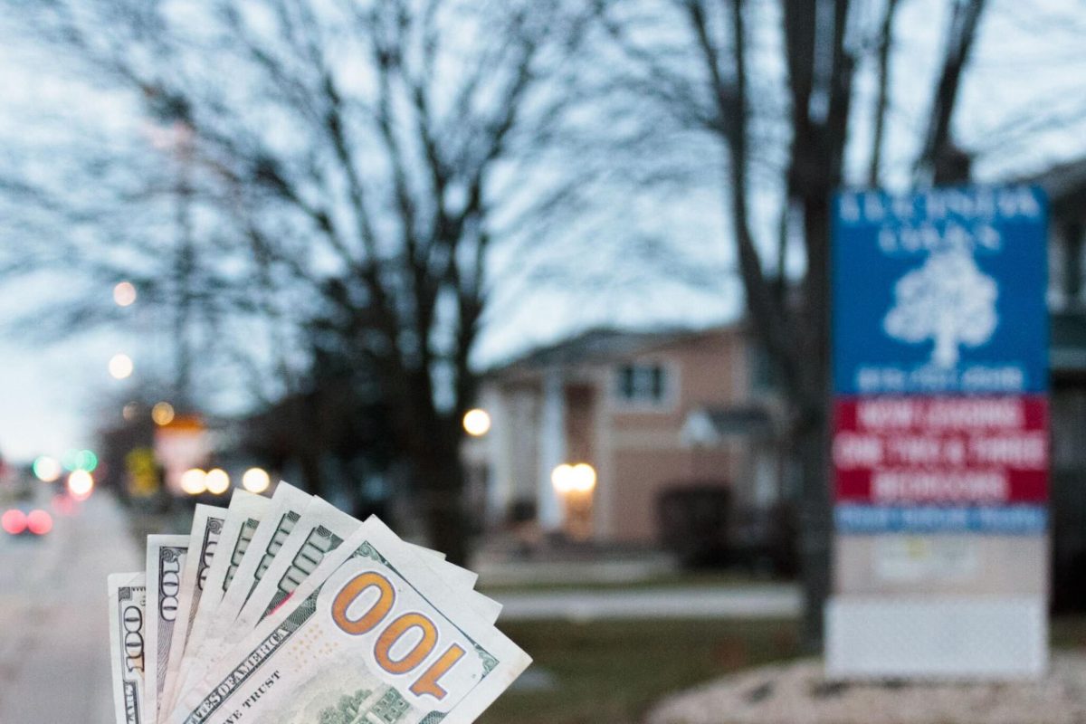 A photo illustration has a hand holding a pile of money in front of a property managers sign. Getting an apartment is expensive, and its important to plan ahead for the expenses that come with an apartment. (Sean Reed | Northern Star)