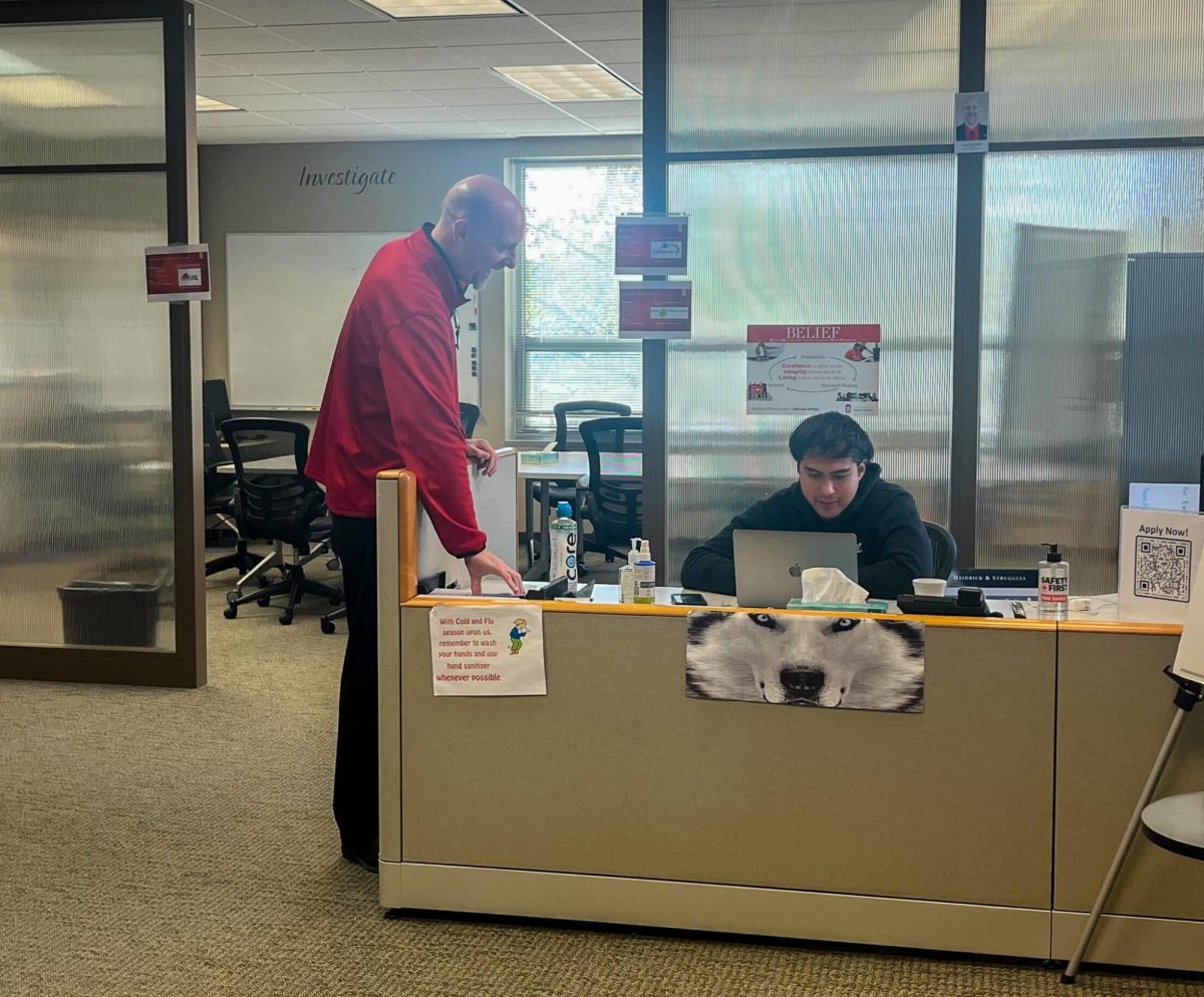 Jose Plascencia, a junior finance major, sits behind a desk, while Jason Gorham, director of Business Consulting in the College of Business, places papers on the desk in the Experiential Learning Center located in Barsema Hall. The Experiential Learning Center provides students with real-world experience working with a company to solve real-world business issues. (Anna Wittenkeller | Northern Star)