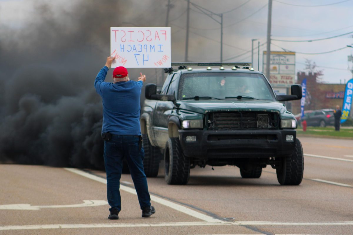 A protester stands on Illinois Route 23 while holding a sign that says “FASCIST IMPEACH 47 NOW” and flips off a truck as it billows smoke from its exhaust pipe. The No Kings protest began just before noon and lasted until around 1:45 p.m. (Ethan Rodriguez | Northern Star)