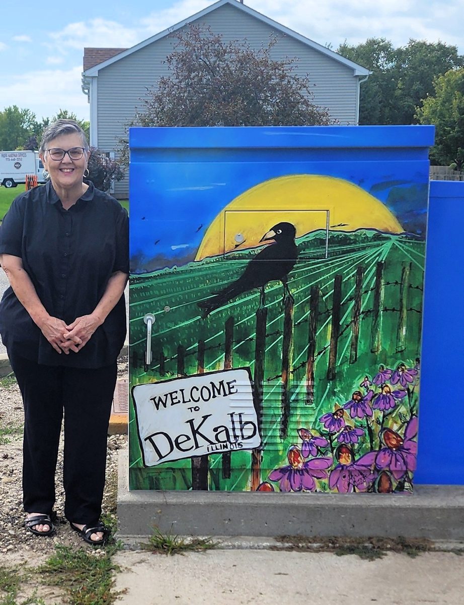 Christine Perkovich of DeKalb stands next to the utility box featuring her painting "Welcome to DeKalb, Illinois" at the entrance to DeKalb High School. Artwork by five DeKalb residents was selected in the fourth round of utility box murals which were recently installed through the public art initiative of the City of DeKalb and its Citizens’ Community Enhancement Commission. (Courtesy of the City of DeKalb and the Citizens’ Community Enhancement Commission)